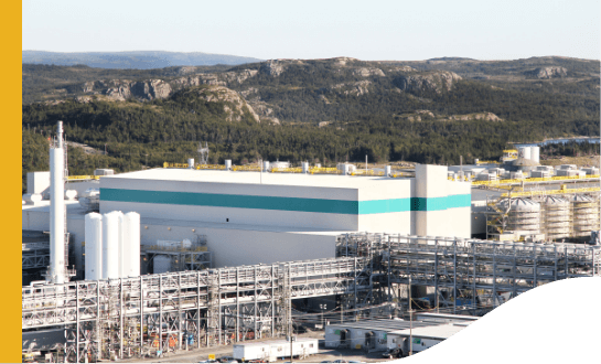 Vista para uma área industrial, com um grande galpão, várias estruturas metálicas e, ao fundo, uma região montanhosa.