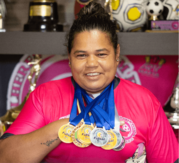 Foto de Lucilene sorrindo para câmera com diversas medalhas em seu pescoço. Atrás dela, há uma estante com prêmios e bolas esportivas.