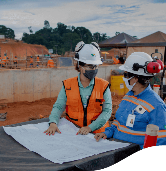 Photo of two women talking and pointing to a big paper, and in the background, an area with construction works with tents, earth, cement and people working. One of the women is wearing a green Vale button-down shirt, an orange vest, goggles, a mask and a helmet. The other woman is wearing a blue shirt with fluorescent lines, a mask, goggles and a helmet.