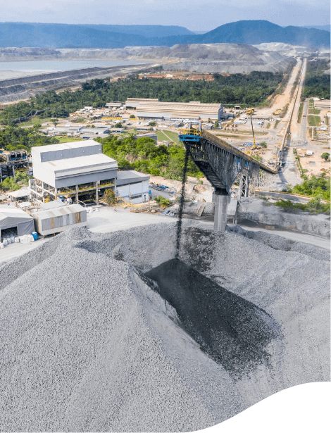 Imagem de uma operação. Em primeiro plano há uma grande pilha de um material em pó cinza e uma estrutura alta despeja um pó preto no mesmo local. Ao fundo é possível ver alguns galpões e uma área montanhosa.