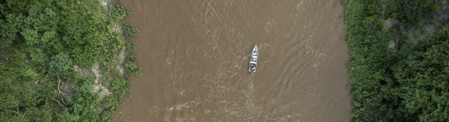 Imagem aérea de um rio de águas turvas com um barco navegando. Aos lados há uma densa vegetação.