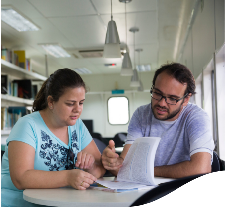 A man and a woman sitting in a library leafing through a book that is propped up on a table