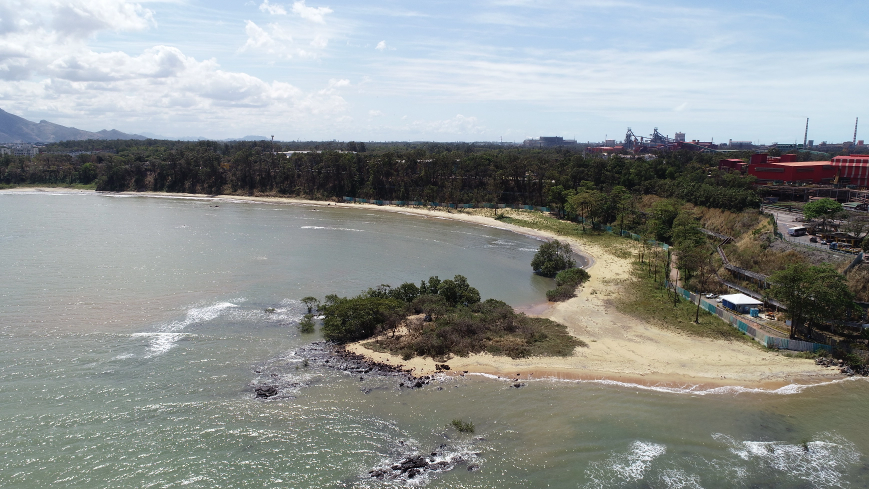 Praia com bastante água à vista, pedras, vegetação viva e areia no solo. Ao redor, mais vegetação viva com árvores grandes, além de construções urbanas e pessoas.