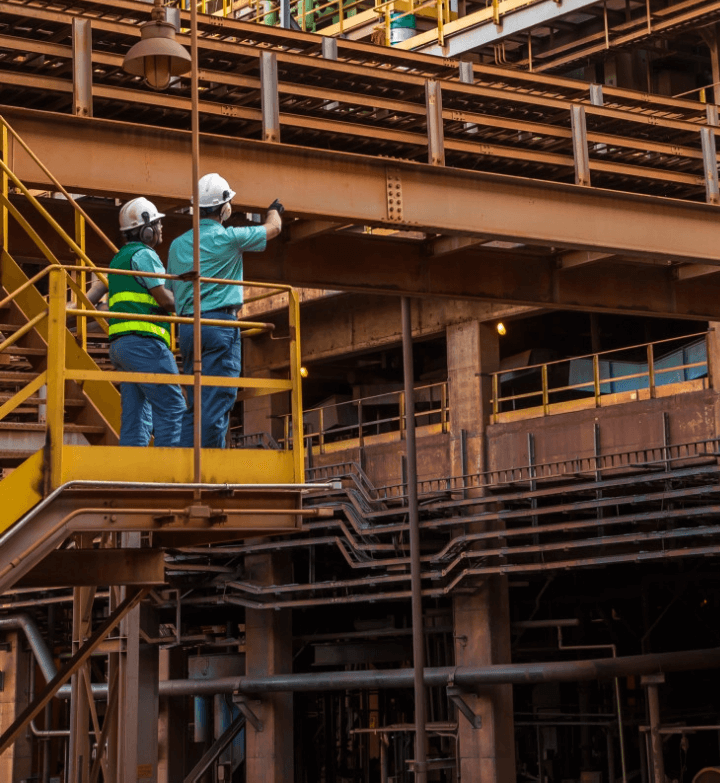 Dois empregados Vale estão sob uma estrutura de ferro e um deles aponta para cima. Ambos estão de capacete de proteção.