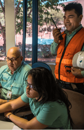 Dois empregados da Vale – um homem e uma mulher – estão sentados lado a lado em uma mesa de escritório e olham para frente. Os dois usam camisas verdes de um tom claro. Atrás deles está um outro empregado, usando camisa verde e colete laranja, em uma das mãos ele segura um capacete branco e na outra um rádio comunicador.