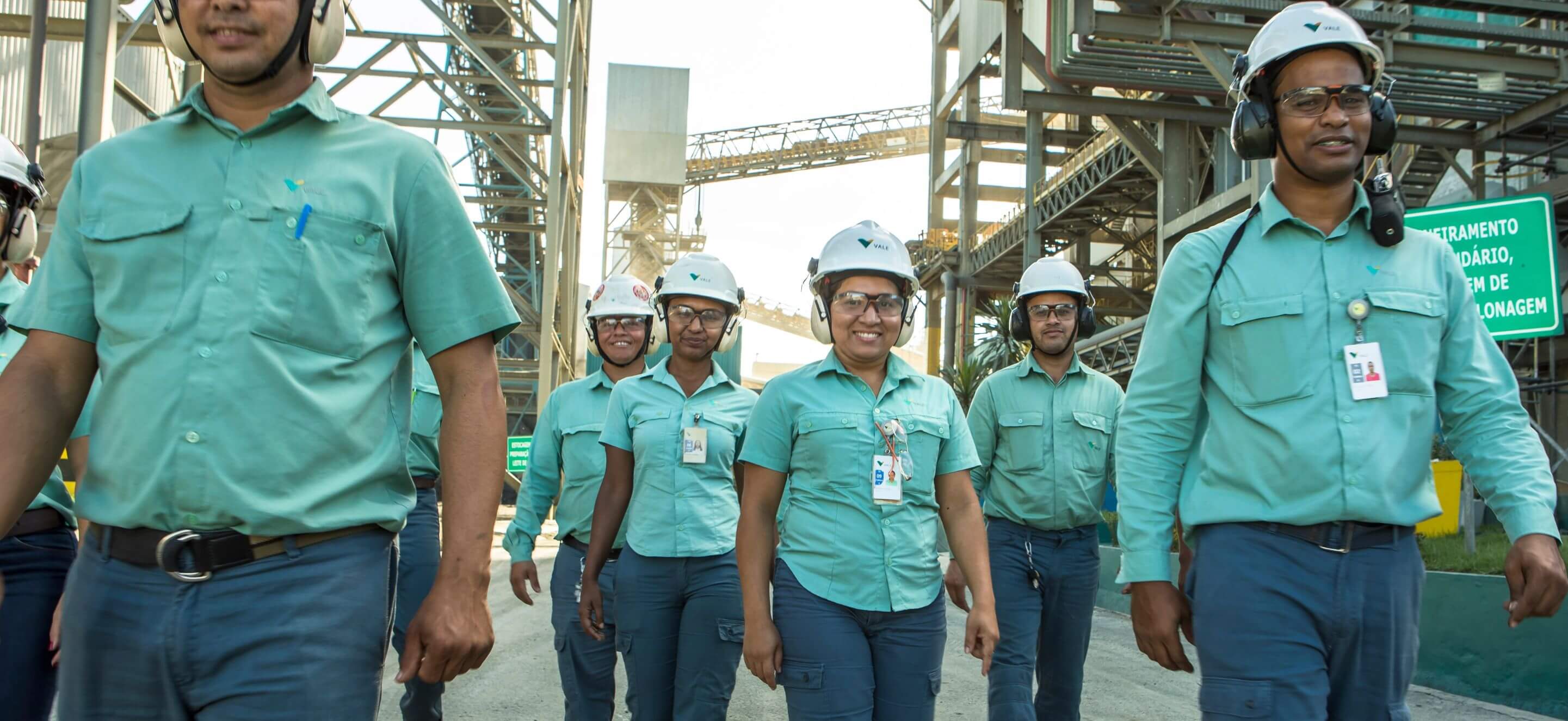 Male and female employees walking in side by side in an operation space. All are wearing green uniforms, goggles, ear muffs and white helmets with Vale logo.