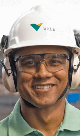 Foto de rosto de um empregado da Vale sorrindo. Ele usa camisa verde clara, óculos de proteção, protetores de ouvido e um capacete branco com logotipo da Vale