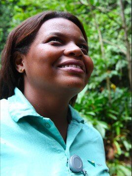 Imagem de busto para cima de uma mulher negra. Ela está sorrindo e usa um uniforme verde claro da Vale. Ao fundo está um espaço arborizado.