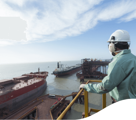 Empregado Vale, vestido com uniforme verde capacete e luvas de proteção, observa um porto com dois navios cargueiros atracados.