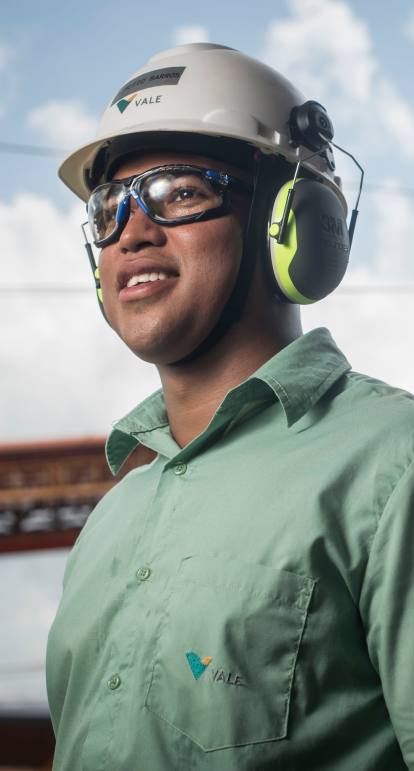 Homem parado ao lado de um trem verde, amarelo e cinza. Ele usa uniforme laranja com logotipo da Vale, óculos de proteção e capacete branco também com logotipo da empresa.