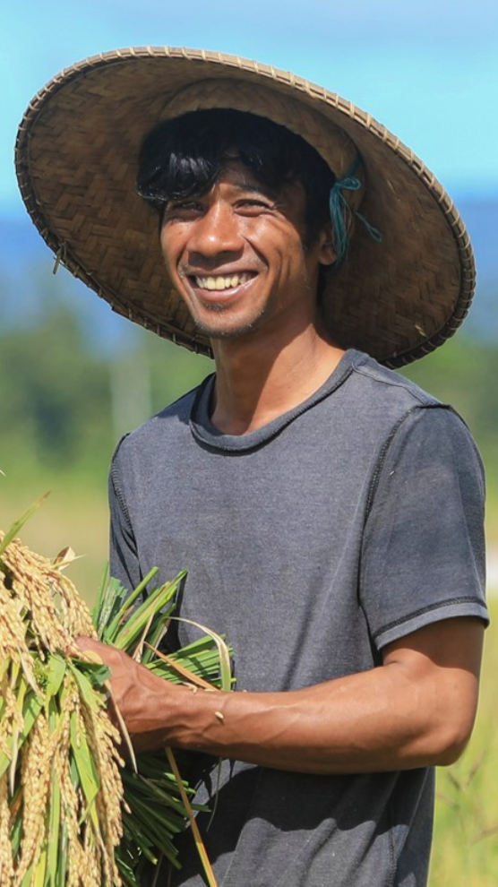 A man in a hat smiles for a photo. In his hands he carries some plants.