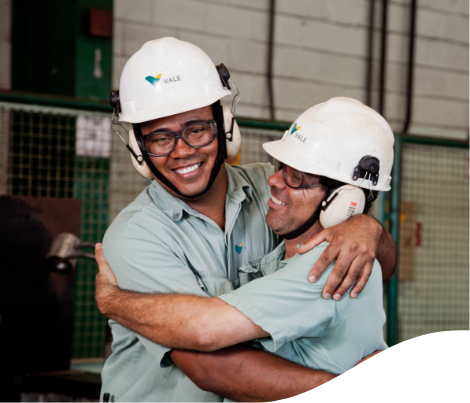 Belly to head shot of two Vale employees hugging and smiling. Both are wearing a Vale uniform, green button-down shirt, ear protection, goggles and helmet.