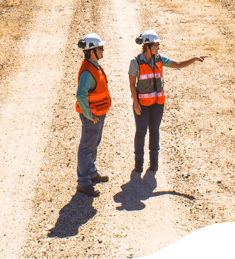 Duas empregadas Vale em um chão de areia. Elas estão de colete laranja e capacetes. Uma aponta para o horizonte e a outra olha na mesma direção.