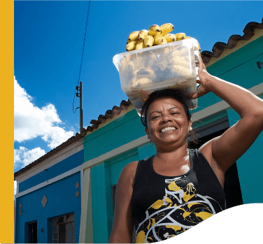 Uma mulher sorri para foto enquanto segura na cabeça uma bacia transparente com bananas. Ao fundo há duas casas azuis de arquitetura antiga.