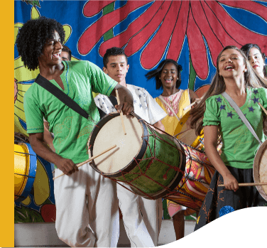 Jovens tocam tambores e sorriem, como se estivessem cantando. Atrás há um tecido estampado com as cores azul, vermelho, amarelo e verde.