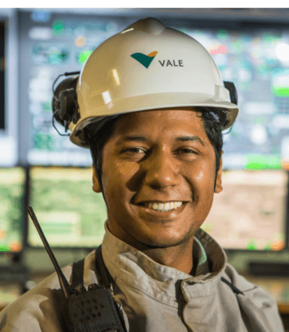 Empregado Vale sorrindo para foto. Ele usa um capacete de proteção e, ao fundo, é possível ver telas de computadores, embaçadas.