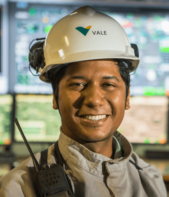 Empregado Vale sorrindo para foto. Ele usa um capacete de proteção e, ao fundo, é possível ver telas de computadores, embaçadas.