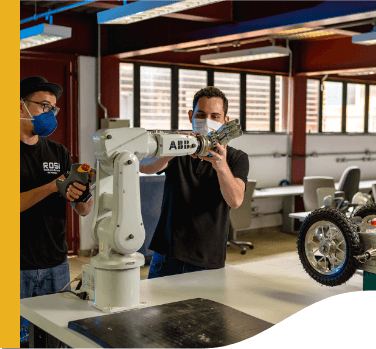 Two men, both wearing black shirts and protective face masks, are working on a robotic prototype that is on a table.