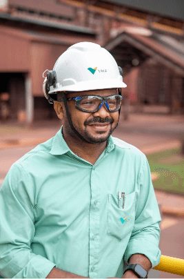 Foto da altura da barriga até a cabeça de um homem de barba sorrindo, usando o uniforme verde da Vale, com capacete e óculos apoiado em uma grade.