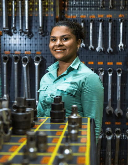 Empregada Vale sorrindo para foto. Ela está vestindo o uniforme verde e, ao fundo, é possível ver ferramentas penduradas na parede.