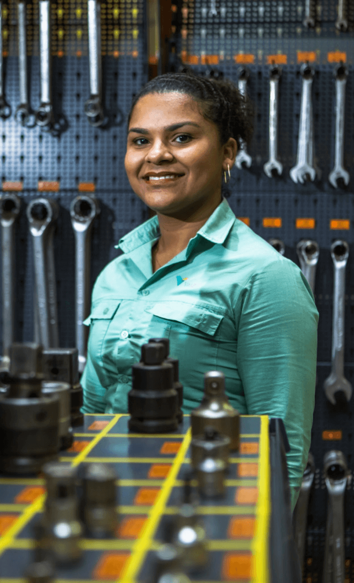 Empregada Vale sorrindo para foto. Ela está vestindo o uniforme verde e, ao fundo, é possível ver ferramentas penduradas na parede.