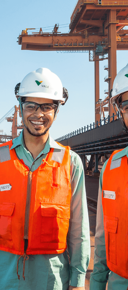 Two men in an operation area near the sea are smiling. They are wearing light green shirts, orange vests, white helmets and goggles