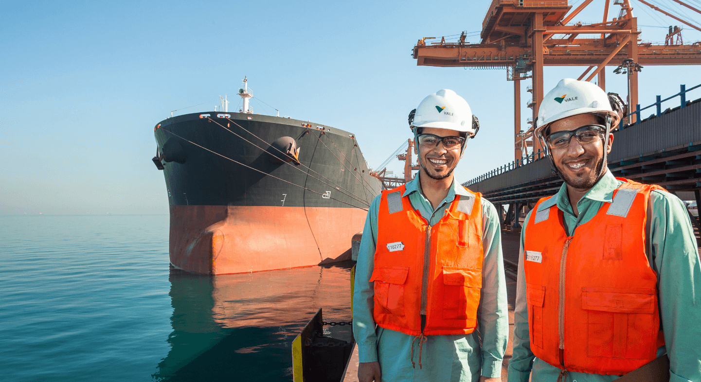 Two men in an operation area near the sea are smiling. They are wearing light green shirts, orange vests, white helmets and goggles