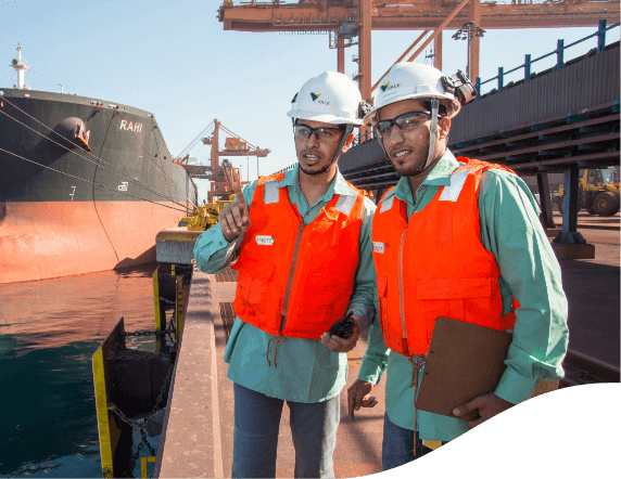 Men standing side by side in a port. A ship is moored next to them and the two look forward to where one of them is pointing. The two wear green shirts, orange vests, goggles and white helmets with Vale logo. One of them holds a clipboard