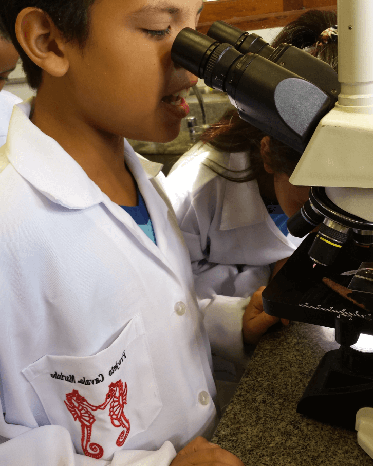 Foto de uma criança olhando um microscópio.  O menino está usando um avental branco