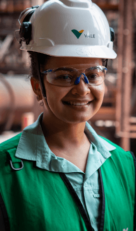 Mulher sorrindo em uma área operacional. Ela usa camisa verde clara, colete em um tom mais escuro de verde, óculos de proteção e capacete branco com logotipo da Vale.