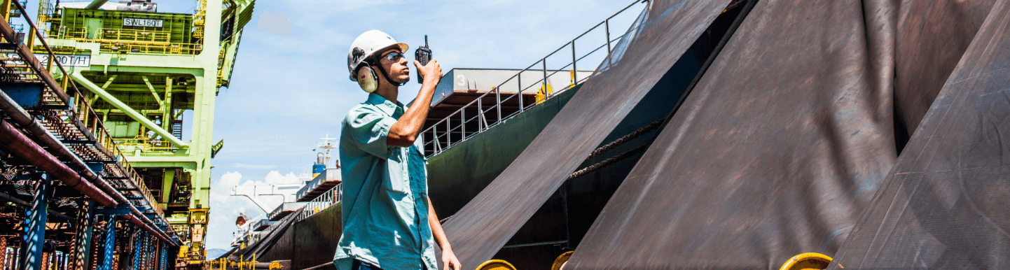 Vale employee standing in the middle of an operations area. He is wearing a light green shirt, goggles, ear muffs, a white helmet with Vale logo and holds a radio communicator close to his mouth with one hand.