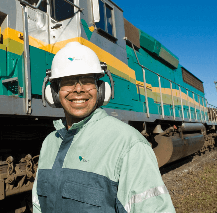 Homem sorrindo ao lado de um trem nas cores verde, amarelo e cinza. O homem usa uniforme verde com logotipo da Vale, óculos de proteção, protetores de ouvido e capacete branco com logotipo de Vale.