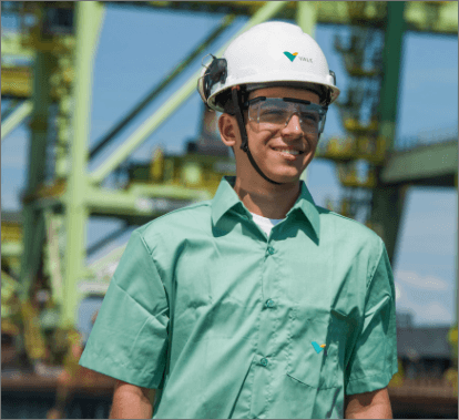 Homem sorrindo em uma área operacional. Ele usa camisa verde clara, óculos de proteção e capacete branco com logotipo da Vale.