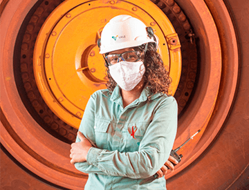 Uma mulher está de braços cruzados e segura um rádio de comunicação em uma das mãos. Ela usa um uniforme verde claro, capacete, óculos e máscara de proteção. Ao fundo está a roda de um caminhão.