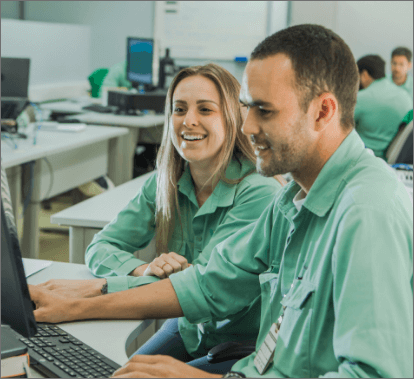Um homem e uma mulher sentados lado a lado em um escritório. Os dois estão usando camisas verdes claras de uniforme e olham para a tela de um computador. A mulher está sorrindo. Ao fundo há outras mesas com computadores e outras pessoas.