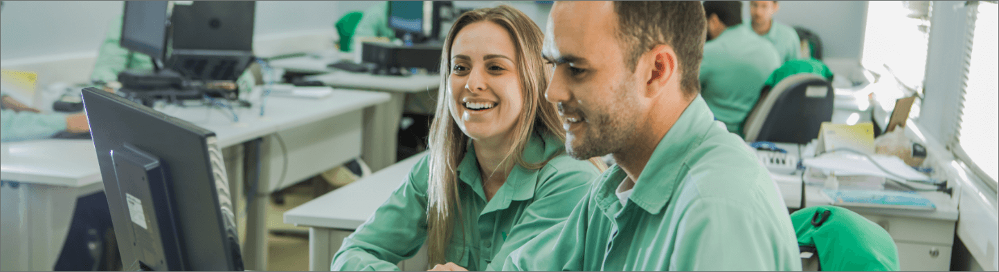 Um homem e uma mulher sentados lado a lado em um escritório. Os dois estão usando camisas verdes claras de uniforme e olham para a tela de um computador. A mulher está sorrindo. Ao fundo há outras mesas com computadores e outras pessoas.