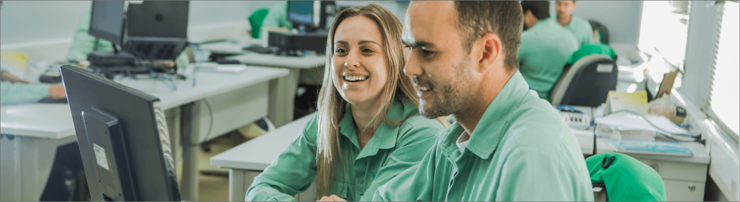 A man and a woman sitting side by side in an office. The two are wearing light green uniform shirts and looking at a computer screen. The woman is smiling. In the background, there are other desks with computers and other people.