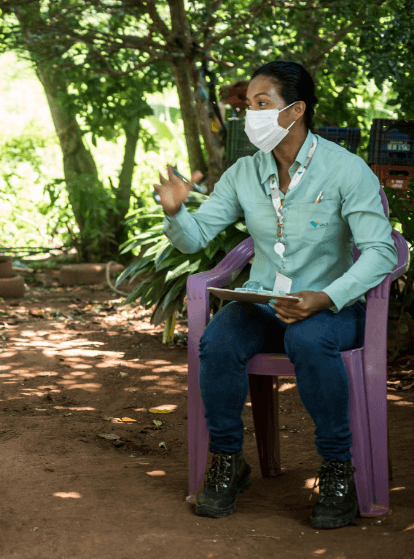 A Vale female employee is sitting on a plastic chair in a dirt yard, with trees in the background. She is gesturing with one hand and holding a clipboard with the other. She is wearing a protective mask as well.