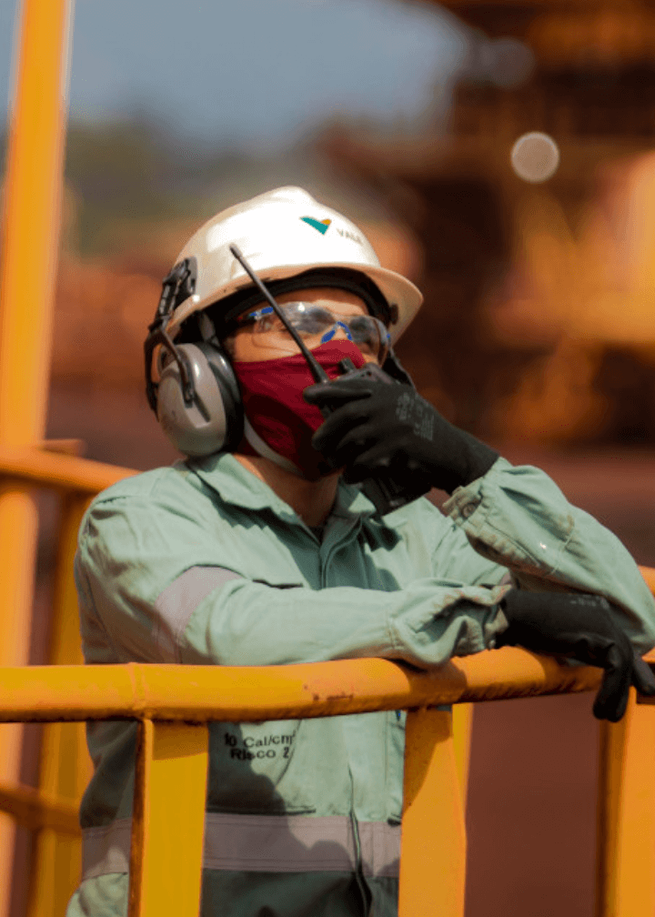 Foto da barriga até a cabeça de uma mulher falando em um rádio em uma operação. A mulher está apoiada em grade e está usando uniforme Vale, capacete, óculos, luva, proteção nos ouvidos e máscara no rosto.