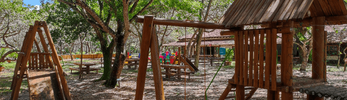 Parquinho infantil ao ar livre. Os brinquedos, como balança e escorregador, são feitos de madeira. Algumas árvores trazem sombra ao local.