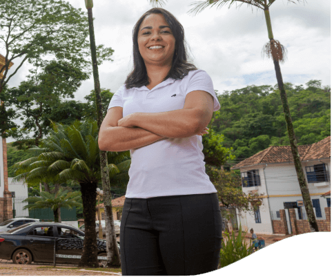 Foto tirada de baixo para cima de mulher com braços cruzados sorrindo para câmera. Ela tem cabelos escuros, lisos e médios. Atrás dela, há uma construção, carros e árvores.