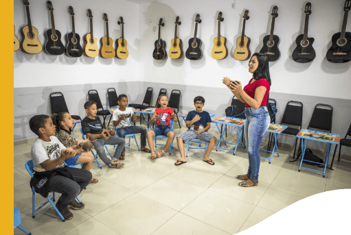 Sala de música com diversos violões pendurados na parede. Ao centro, há diversas crianças sentadas em círculos prestando atenção em uma mulher que gesticula.