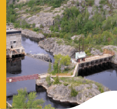 Photo of Wabageshik small hydropower plant with a concrete structure, rocks, vegetation, and moving water passing through the structures.