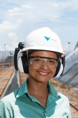 there is female employee, outdoors in an operation. She is wearing a helmet and goggles, smiling, and wearing a green uniform.