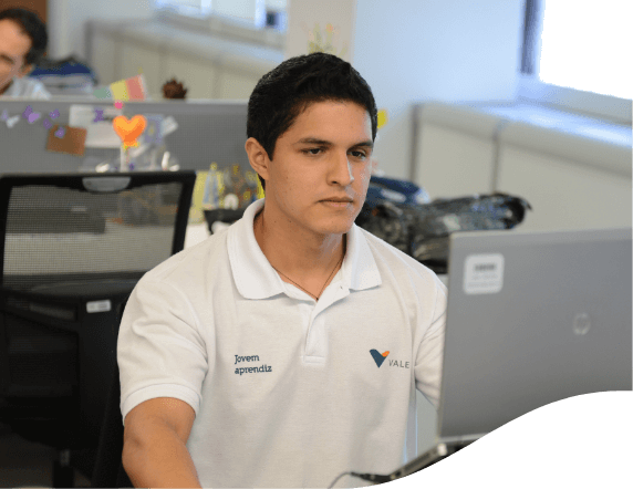 A man sitting in an office looking at a laptop in front of him. He is wearing a white T-shirt with Vale logo.