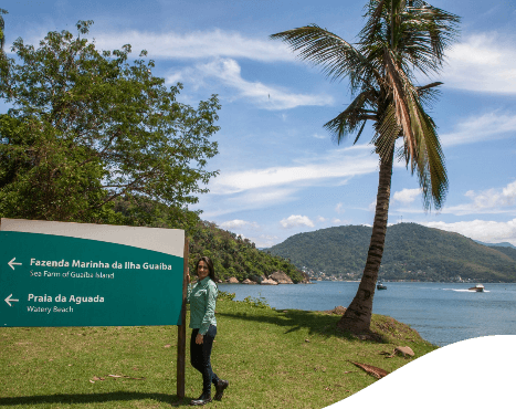 Mulher usando calça jeans e camisa verde clara da Vale está parada ao lado de uma placa onde se lê “Fazenda Marinha da Ilha Guaíba” e “Praia da aguada”. Ao fundo está um espaço com árvores e um lago.