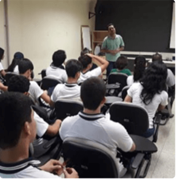 Diversas pessoas estão sentadas, de costas para foto, em uma sala de aula. Ao fundo, de pé, há um empregado Vale.