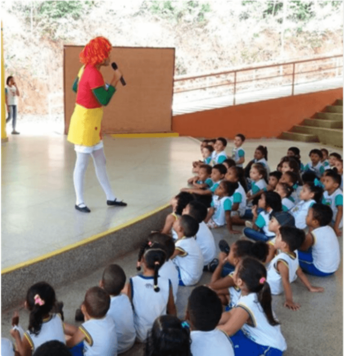 On a stage, a woman is dressed as a rag doll and speaks in a microphone. Several children in school uniforms are sitting on the floor watching the presentation.