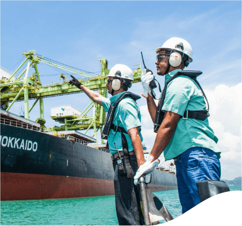 Dois empregados da Vale de frente para um navio em uma área operacional. Os dois usam camisa verde, equipamentos de proteção presos ao corpo, óculos de proteção, protetores de ouvido e capacete branco.