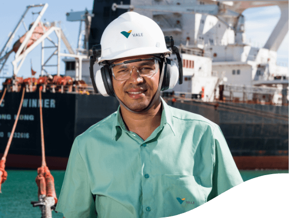 Empregado Vale de uniforme verde, capacete e óculos de proteção. Ele olha para foto e sorri timidamente. Ao fundo é possível ver um navio atracado.